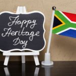 Small sign on small easel painted white next to small south african flag. the sign reads Happy Heritage Day