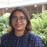 Lady wearing navy blue blouse and glasses standing in front of greenery