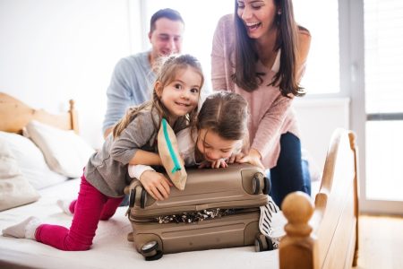 Family on a bed packing a suitcase which is overflowing and the children are trying to foce it closed as Mum helps and Dad looks on. Used in a blog with a list of 10 tips to help with planning a family holiday