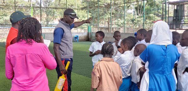A man with a cricket bat showing a group of children with their backs to the camera how to play cricket