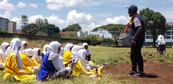 A group of girls sitting on a cricket pitch listening to a man talk.